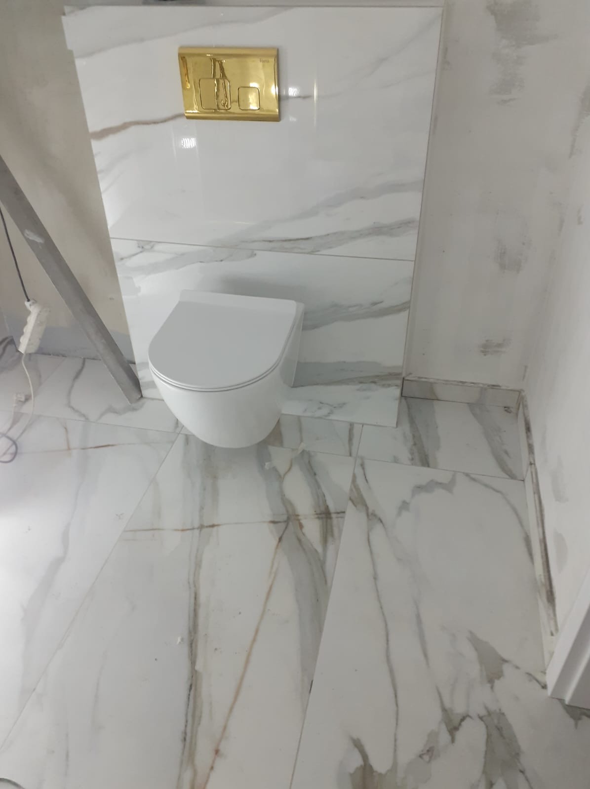 Wisząca toaleta z białej ceramiki, zamontowana na ścianie wyłożonej płytkami imitującymi marmur, z widocznym złotym przyciskiem spłuczki i fragmentem drabiny malarskiej.