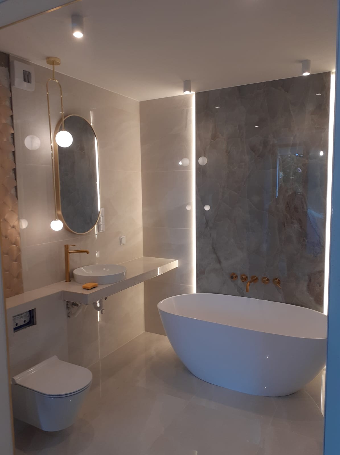 Nowoczesna łazienka z owalną wanną, umywalką nablatową z armaturą w kolorze złotym, podświetlanym lustrem i płytkami imitującymi marmur na ścianie.