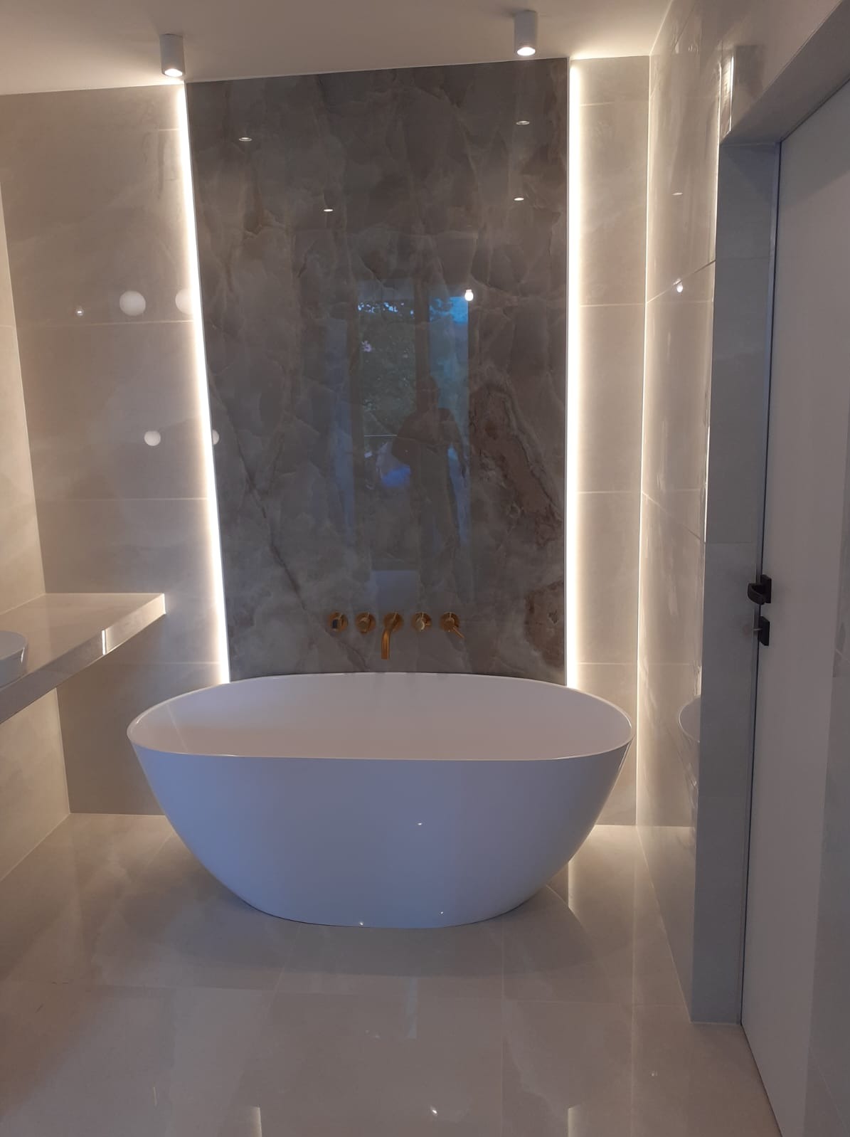 Nowoczesna łazienka z wolnostojącą białą wanną, złotą armaturą ścienną i podświetlanymi panelami z imitacją marmuru.