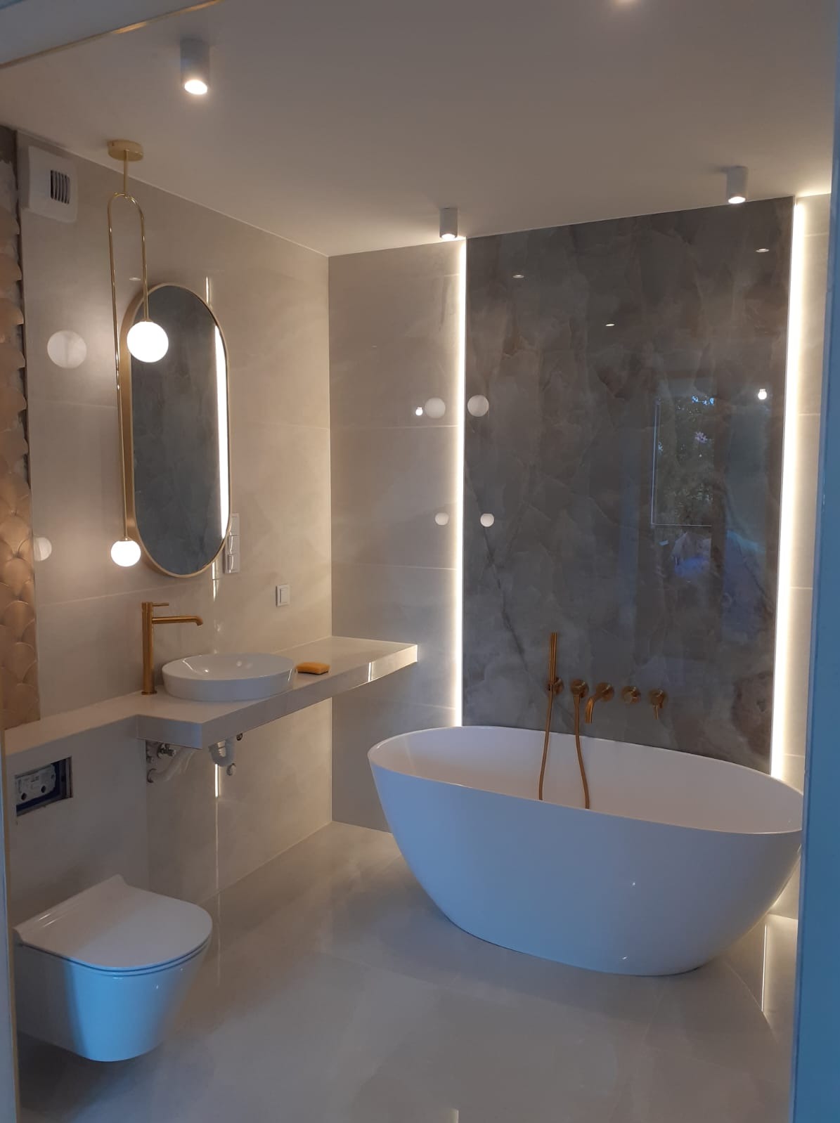 Nowoczesna łazienka z owalną wanną wolnostojącą, umywalką nablatową z złotą armaturą, podświetlanym lustrem i płytkami imitującymi marmur.