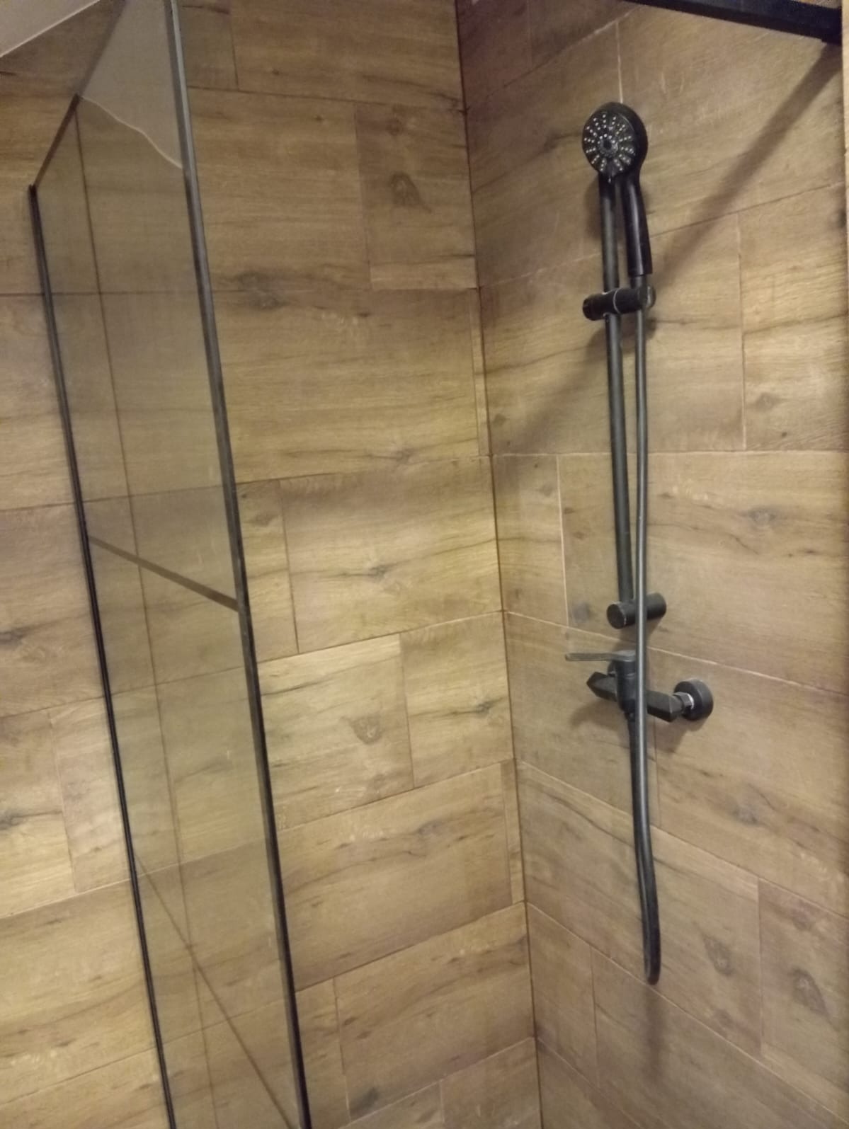 Kabina prysznicowa z czarną armaturą na ścianie wyłożonej płytkami imitującymi drewno.