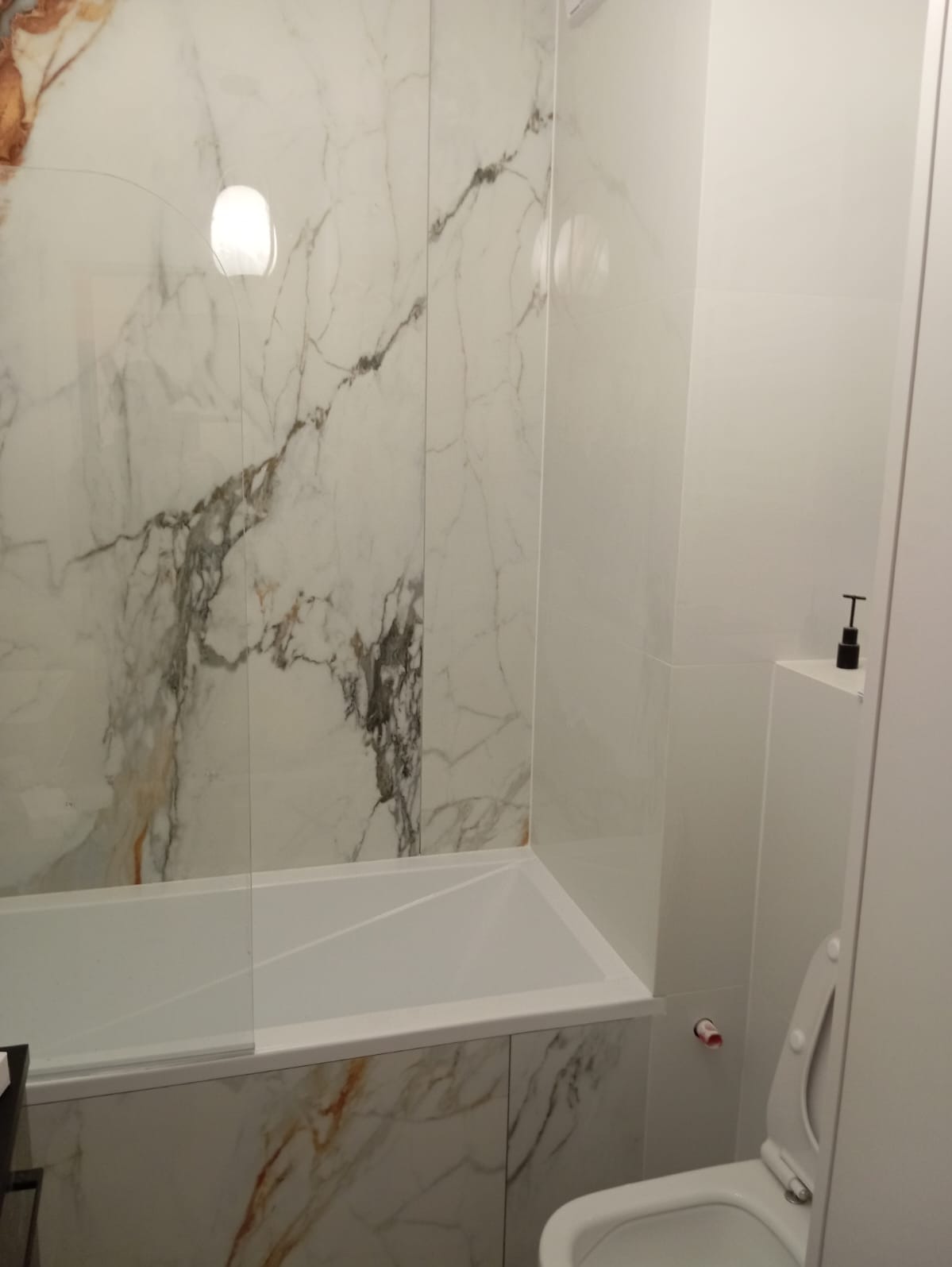 Nowoczesna łazienka z wanną obudowaną płytkami imitującymi marmur, szklany parawan nawannowy, biała toaleta i dozownik mydła na półce.