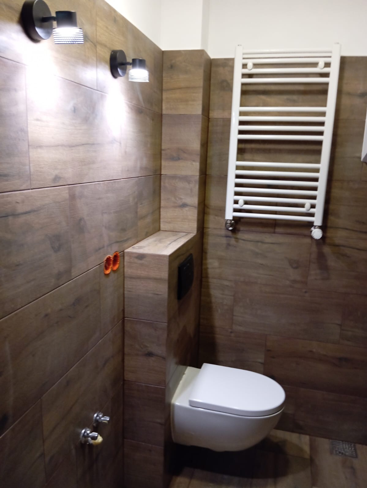 Nowoczesna łazienka z brązowymi płytkami imitującymi drewno, wisząca toaleta, biały grzejnik drabinkowy, dwa czarne kinkiety z białymi kloszami i pomarańczowe zaślepki instalacji wodnej.