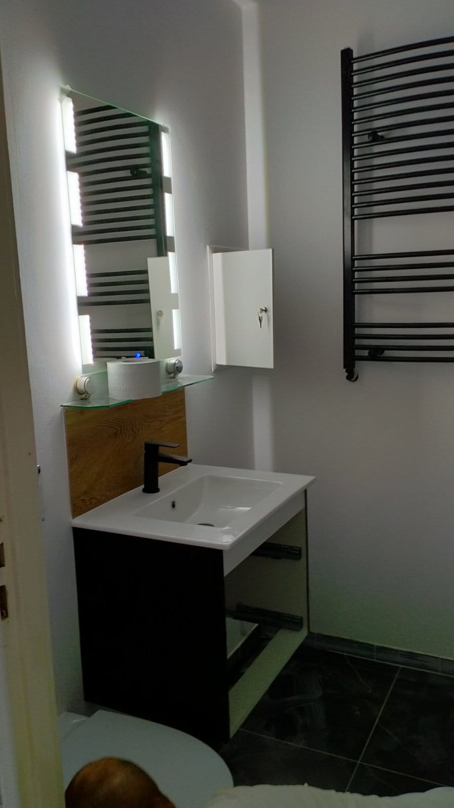 Nowoczesna łazienka z umywalką nablatową, czarną armaturą i podświetlanym lustrem, uzupełniona o grzejnik drabinkowy i szafkę z kluczem.