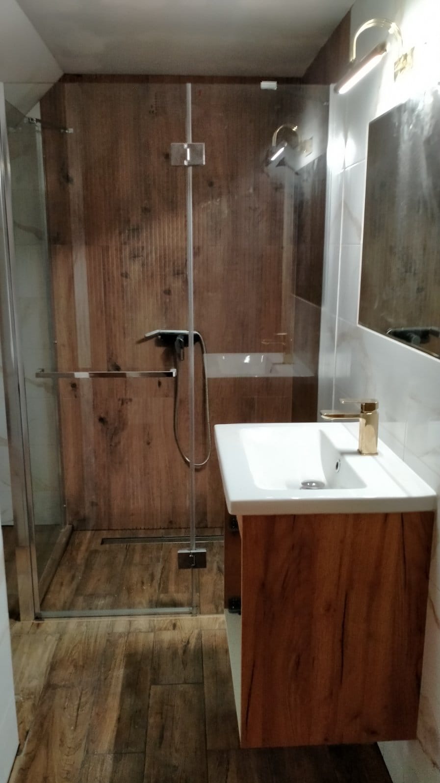 Ukończona łazienka z kabiną prysznicową z transparentnego szkła z chromowanymi okuciami, umywalką nablatową na szafce z drewnopodobnym fornirem i złotą baterią, wykończona płytkami imitującymi drewno.
