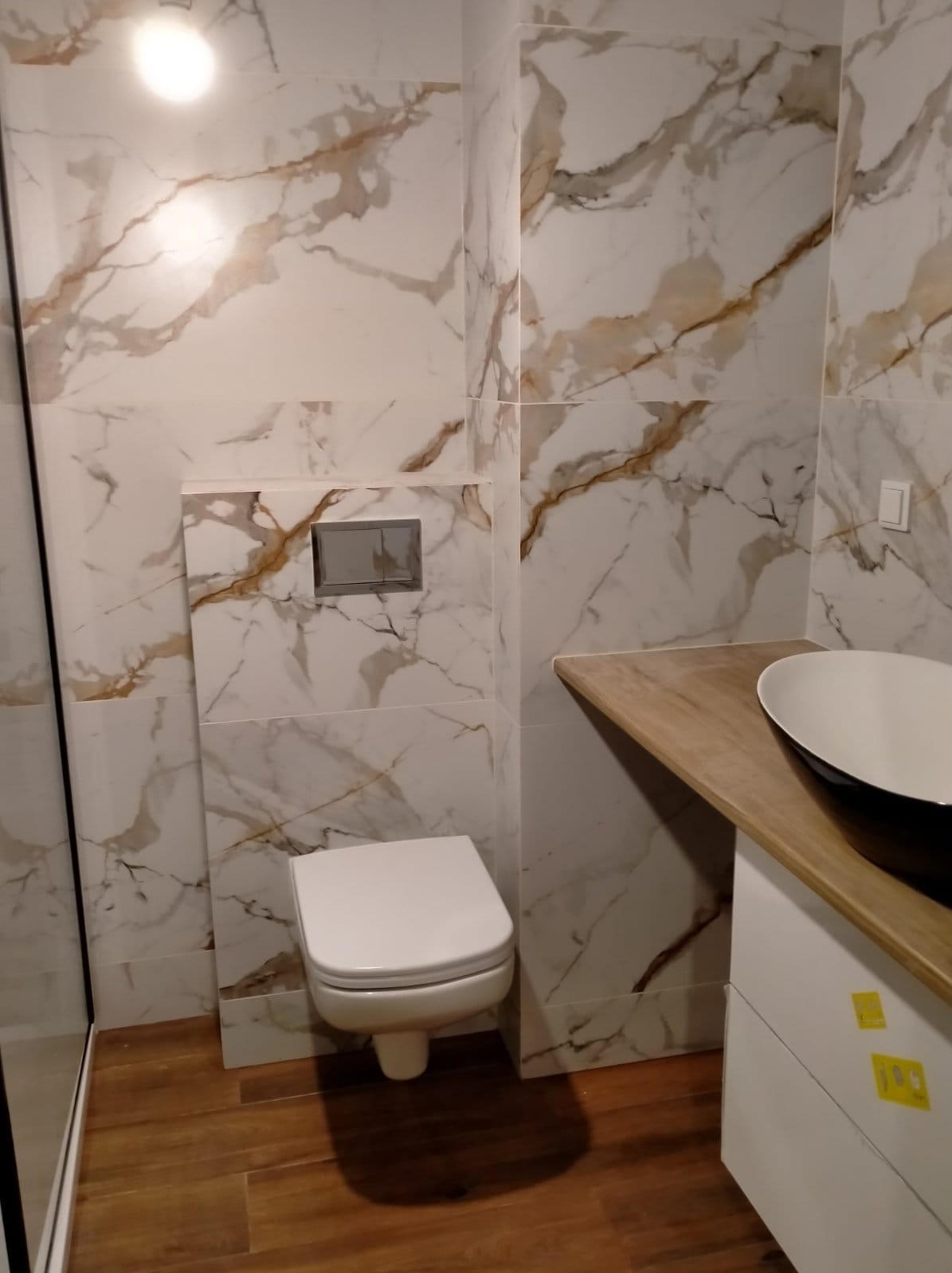 Nowoczesna łazienka z marmurowymi płytkami, podwieszana toaleta z chromowanym przyciskiem spłukiwania i umywalka nablatowa w czarno-białej kolorystyce na drewnianym blacie.