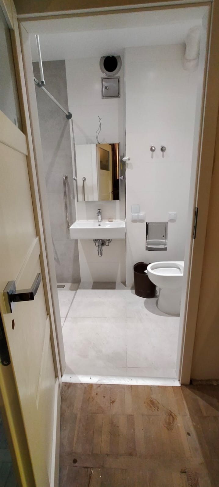 Widok na wykończoną łazienkę z białymi płytkami, umywalką z szafką, toaletą i brązowym koszem na śmieci, widoczny przez otwarte drzwi z drewnianą podłogą przed wejściem.