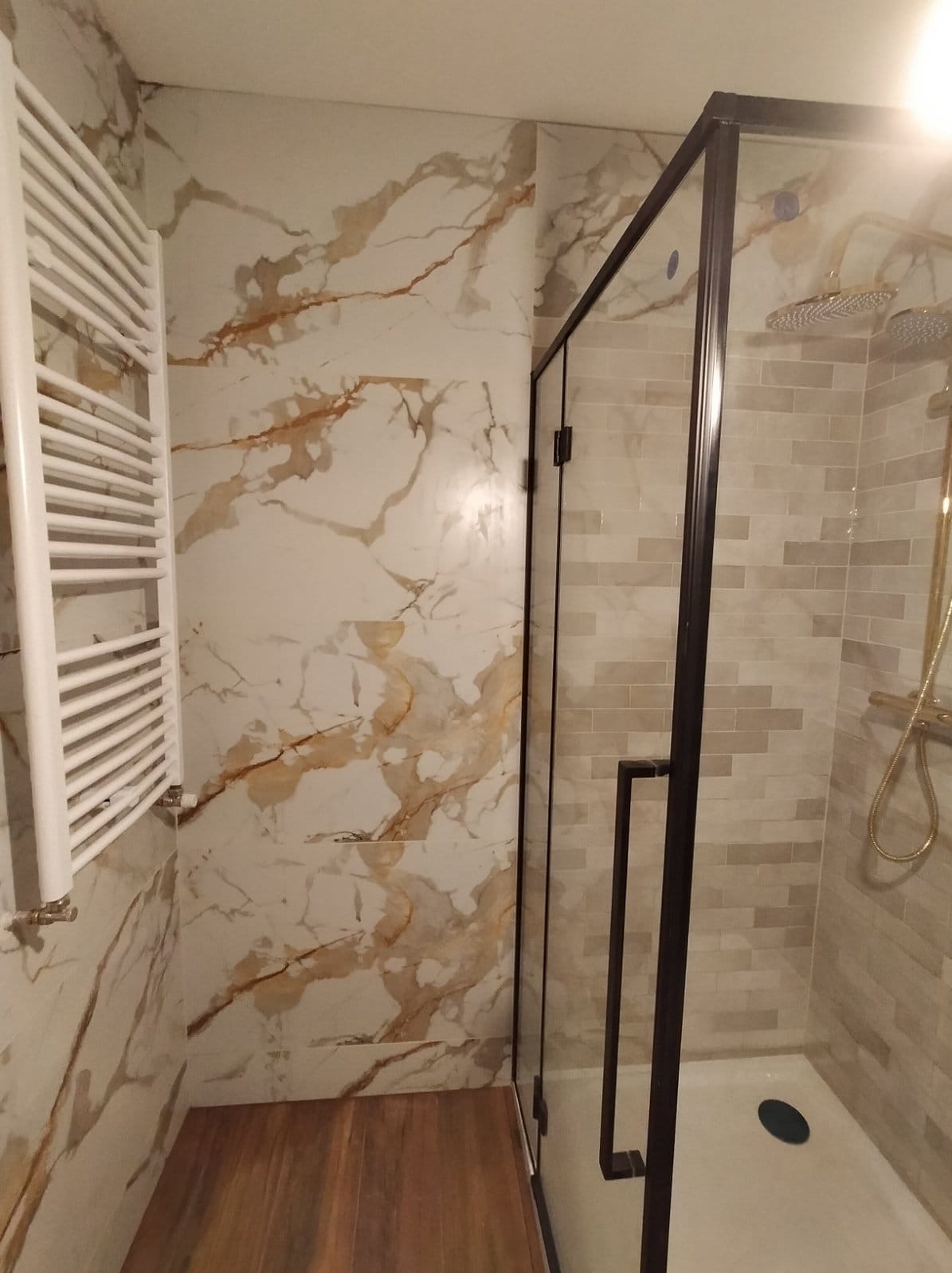 Nowoczesna łazienka po remoncie: kabina prysznicowa z czarną ramą, płytki imitujące marmur z brązowymi żyłami na ścianie, grzejnik drabinkowy i drewnopodobna podłoga.