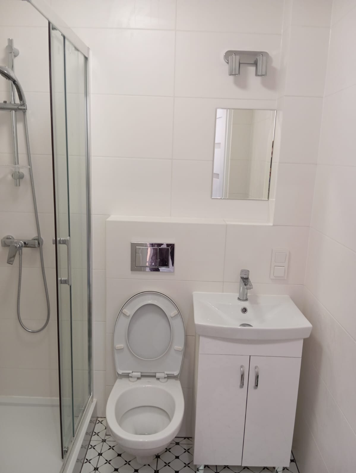 Biała, nowoczesna łazienka z kabiną prysznicową, toaletą podwieszaną z chromowanym przyciskiem spłukiwania, umywalką z szafką i lustrem, wykończona białymi płytkami i wzorzystą podłogą.