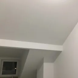 Białe wnętrze poddasza z oknem, widoczne gładkie ściany i sufit z elementami konstrukcyjnymi, pomalowane na biało.