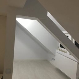 Private - Jasne poddasze po remoncie, z białymi ścianami, podłogą w odcieniach szarości i skośnym sufitem z widocznymi belkami konstrukcyjnymi, grzejnik pod oknem.