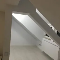 Jasne poddasze po remoncie, z białymi ścianami, podłogą w odcieniach szarości i skośnym sufitem z widocznymi belkami konstrukcyjnymi, grzejnik pod oknem.