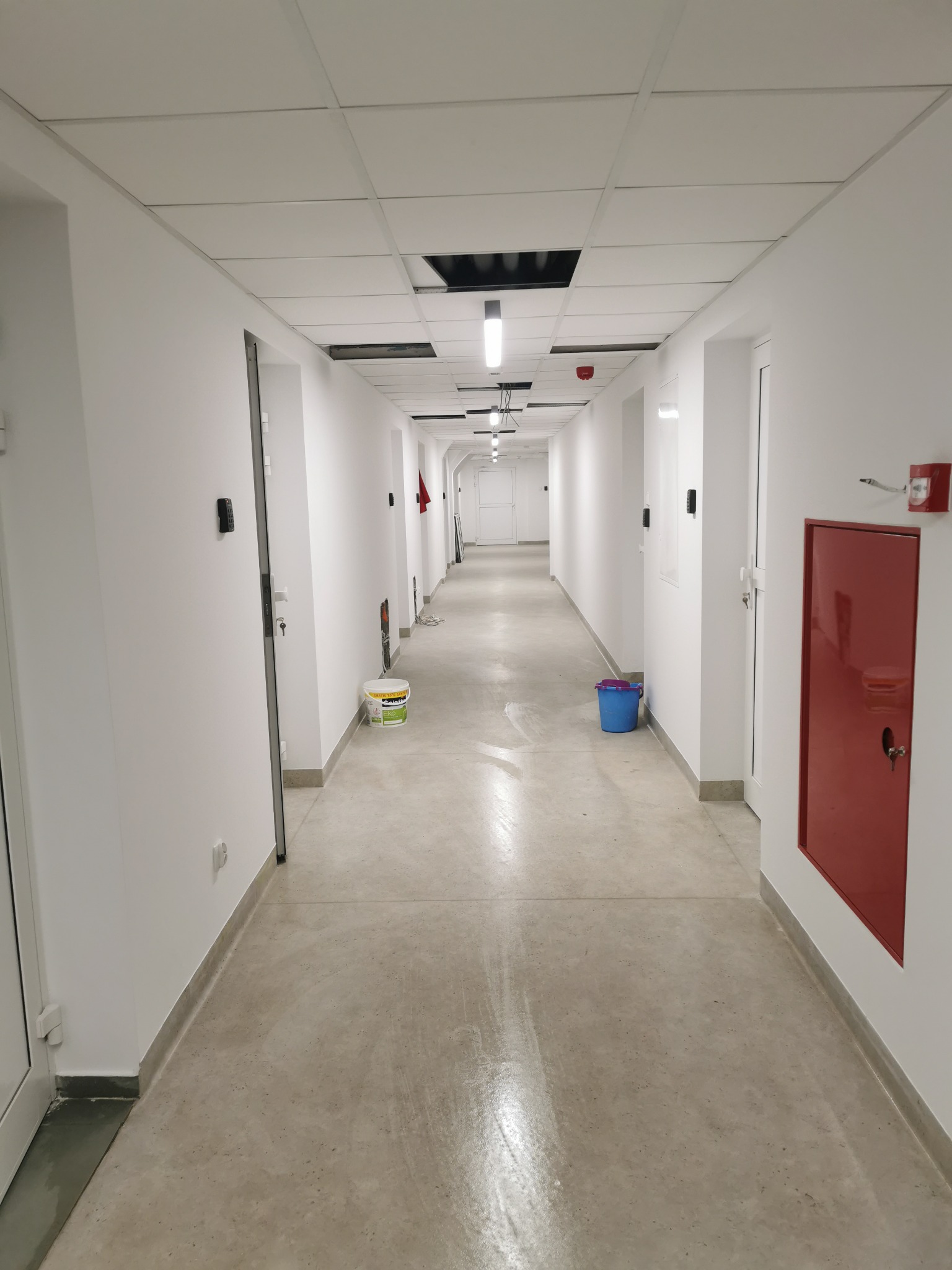 Długi korytarz biurowy w Gdyni po remoncie, z białymi ścianami, szarą podłogą, białym sufitem podwieszanym z widocznymi otworami instalacyjnymi, wiadrem i pojemnikiem po farbie w tle.
