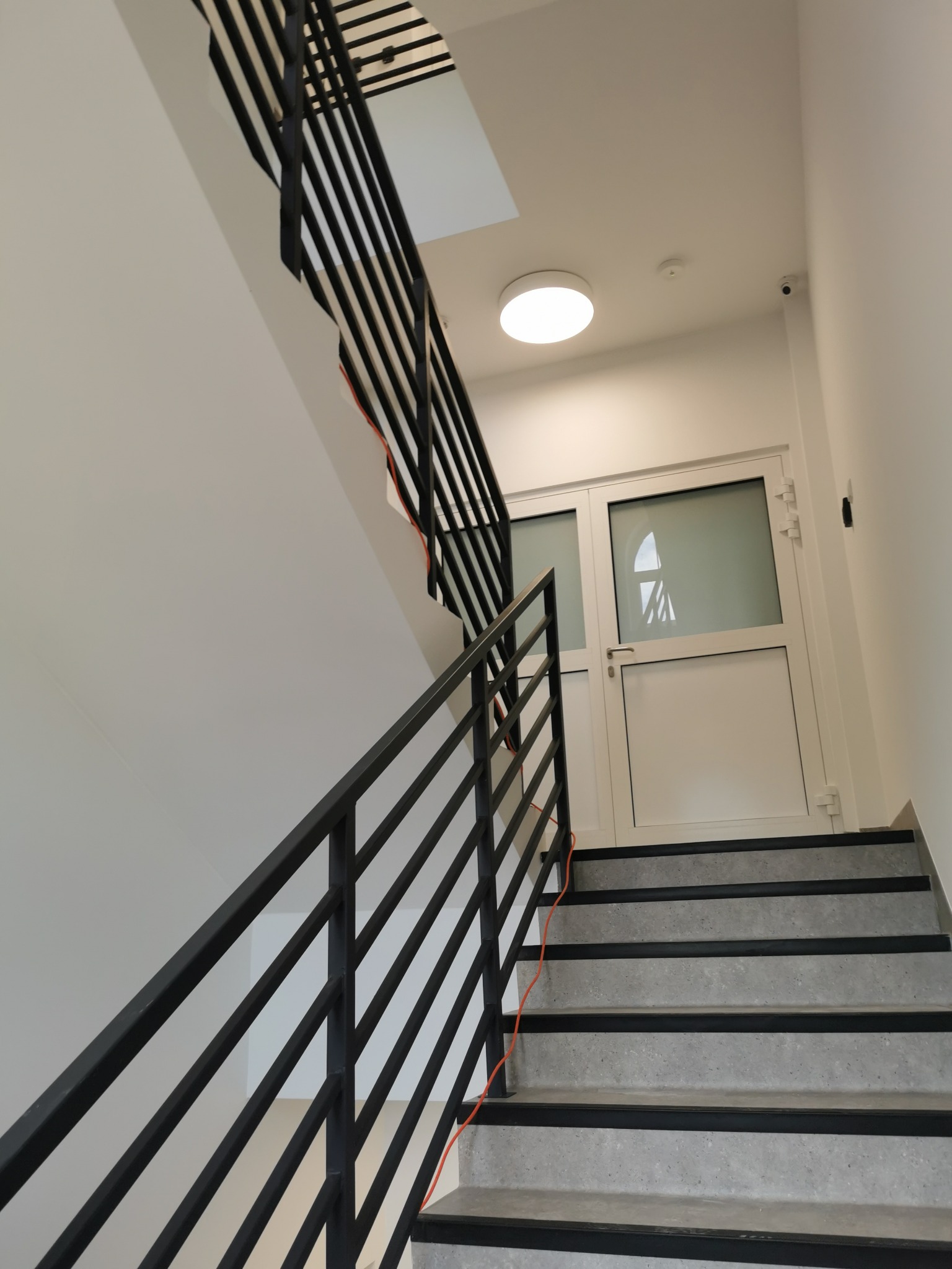 Klatka schodowa w jasnej kolorystyce z czarnymi elementami: balustrada, pasy antypoślizgowe na stopniach, widoczne okablowanie elektryczne w rogu.