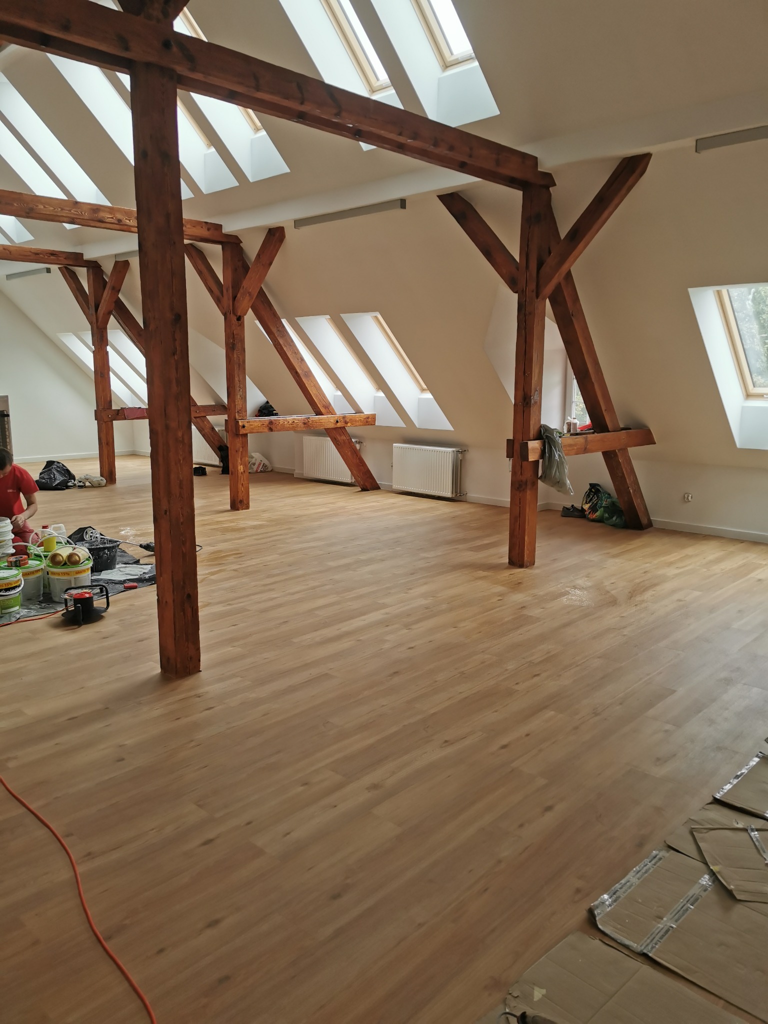 Przestronne poddasze z drewnianymi belkami konstrukcyjnymi i oknami dachowymi w trakcie prac wykończeniowych, widoczne puste ściany przygotowane do malowania oraz akcesoria malarskie.