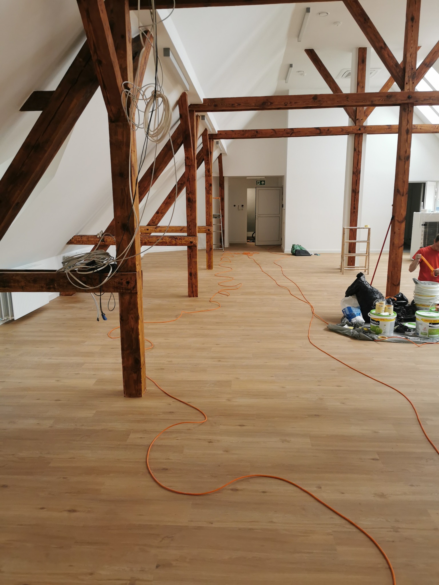 Przestronne poddasze z drewnianą konstrukcją, przygotowane do malowania: widoczne wiadra z farbą, folia ochronna i rozciągnięty pomarańczowy kabel zasilający.
