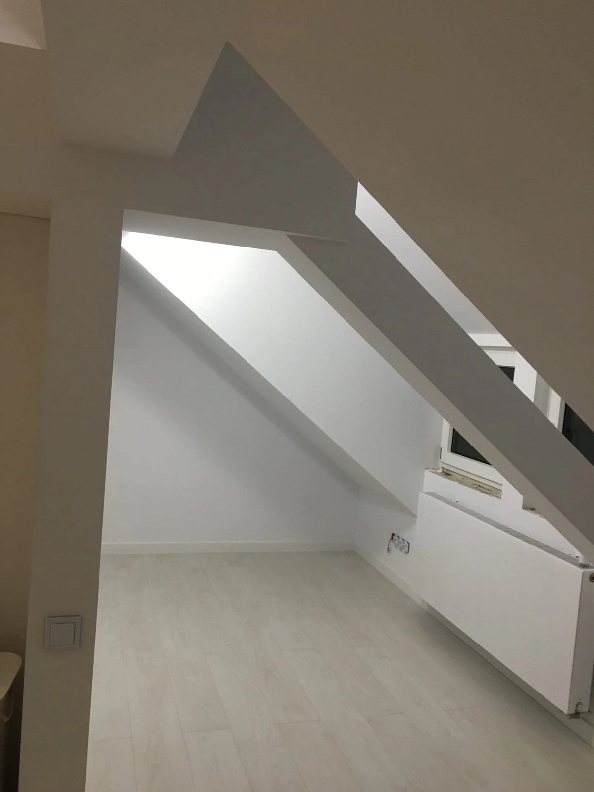 Jasne poddasze po remoncie, z białymi ścianami, podłogą w odcieniach szarości i skośnym sufitem z widocznymi belkami konstrukcyjnymi, grzejnik pod oknem.