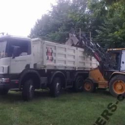 Żółta koparko-ładowarka nabiera ziemię do białej ciężarówki z czerwoną grafiką na pace. Teren trawiasty, drzewa w tle. Dzień pochmurny.