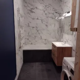 Nowoczesna łazienka z marmurowymi płytkami, drewnianą szafką pod umywalką, grzejnikiem drabinkowym i pralką Whirlpool w rogu.