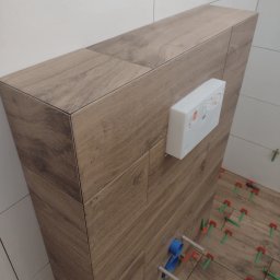 Andrzej Remont - Stelaż podtynkowy obudowany płytkami imitującymi drewno, z widocznym przyciskiem spłukującym i systemem poziomowania płytek na podłodze, w trakcie prac wykończeniowych.