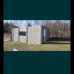 Surowy, betonowy płot modułowy z czarnym panelem wbudowanym, na trawiastym terenie, drzewa w tle.