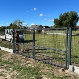 Montaż dwuskrzydłowej bramy wjazdowej z siatki metalowej, widoczny pracownik z narzędziami i samochód dostawczy na tle błękitnego nieba i domów.