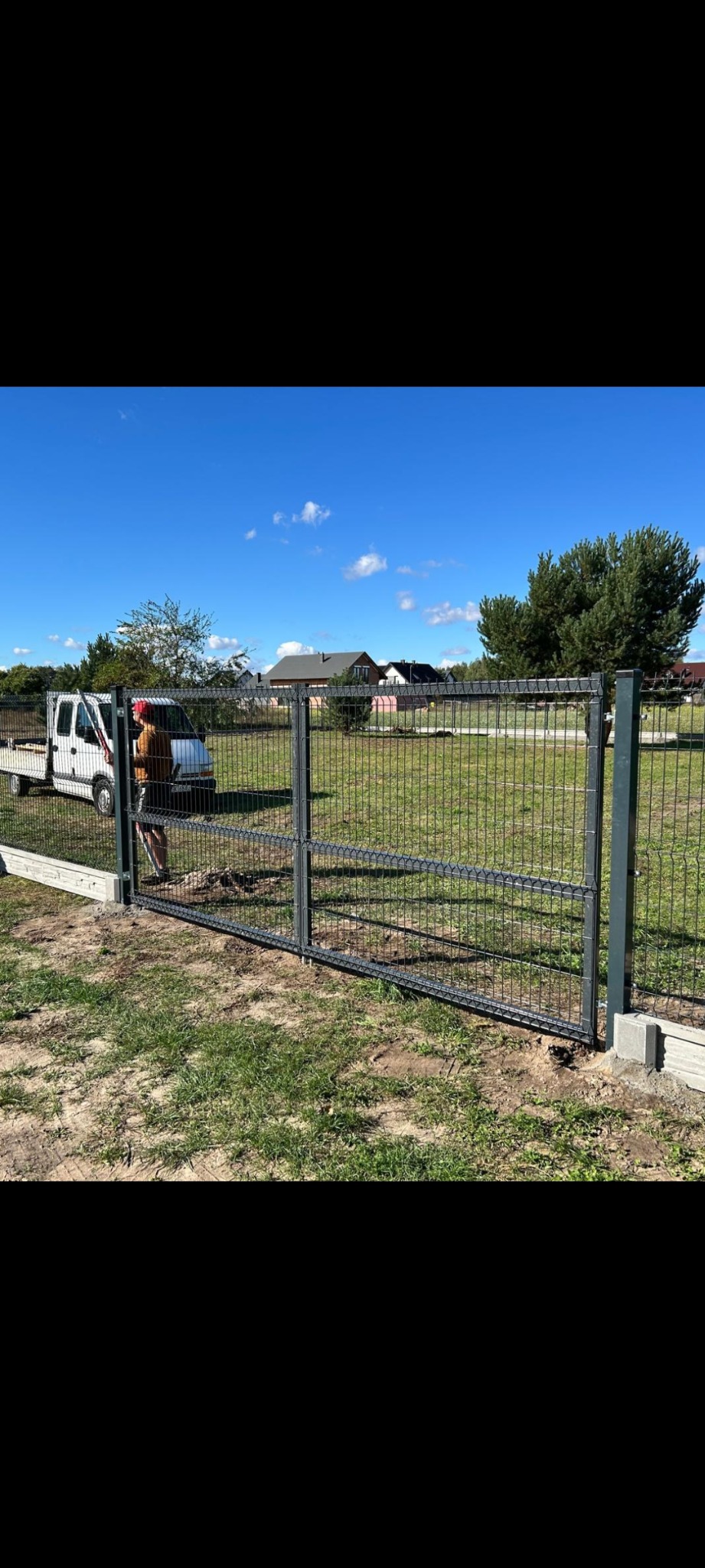 Montaż dwuskrzydłowej bramy wjazdowej z siatki metalowej, widoczny pracownik z narzędziami i samochód dostawczy na tle błękitnego nieba i domów.