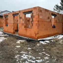 Wznoszone ściany domu murowanego z czerwonej ceramiki poryzowanej, widoczne czarne elementy nadproży okiennych, na ziemi błoto i resztki śniegu, w tle drugi budynek z ciemną dachówką.