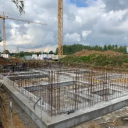 Zalany betonowy fundament domu z wystającymi prętami zbrojeniowymi, widoczne dwa żółte dźwigi budowlane w tle.