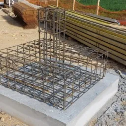 Klatka zbrojeniowa na betonowym fundamencie, widoczne szalunki i teren budowy.