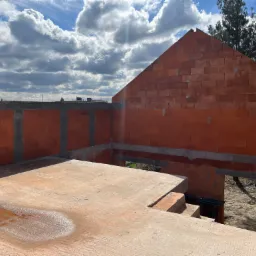 Surowy stan budynku z czerwonej cegły, widoczny z g&oacute;ry, z mokrą wylewką na podłodze, na tle pochmurnego nieba i drzew.