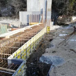 Budowa betonowej konstrukcji oporowej z użyciem szalunków systemowych Doka i zbrojenia stalowego, na tle zalesionego zbocza.