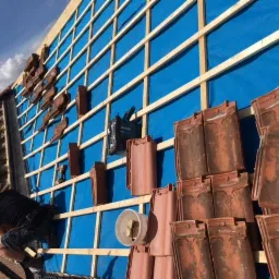 Pracownik montuje dachówkę na drewnianej konstrukcji pokrytej niebieską membraną, widoczne narzędzia i stos dachówek.