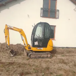 Żółta mini koparka JCB 8018 na placu budowy przed białym domem z drewnianą konstrukcją werandy i balkonem, obok hałda ziemi.