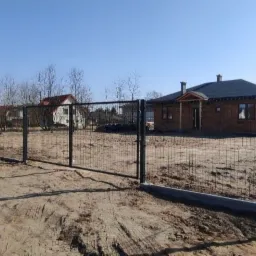 Nowoczesne ogrodzenie panelowe z podmurówką betonową w kolorze grafitowym, otaczające plac budowy domu jednorodzinnego w słoneczny dzień.