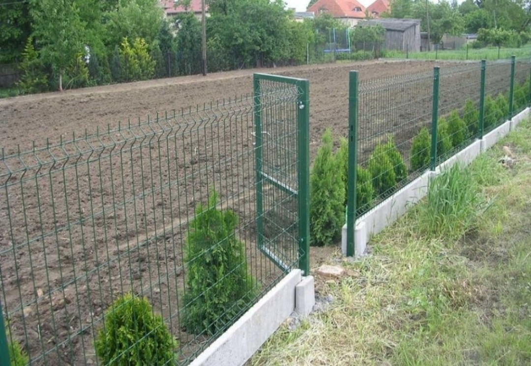 Zielone ogrodzenie panelowe z furtką, osadzone na betonowej podmurówce, z nasadzonymi tujami wzdłuż ogrodzenia, widok na zaorane pole i zabudowania w tle.