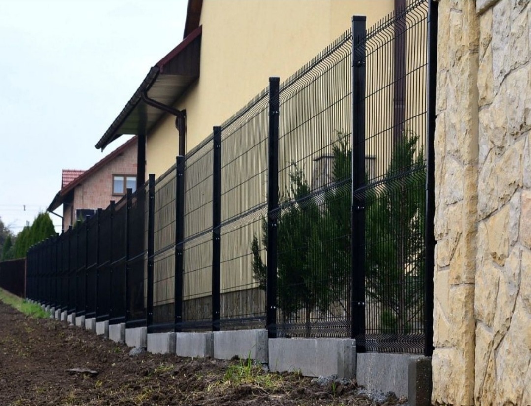 Długi, czarny ogrodzenie panelowe na betonowej podmurówce, z widocznymi drzewami iglastymi za nim i kamiennym słupkiem z prawej strony.