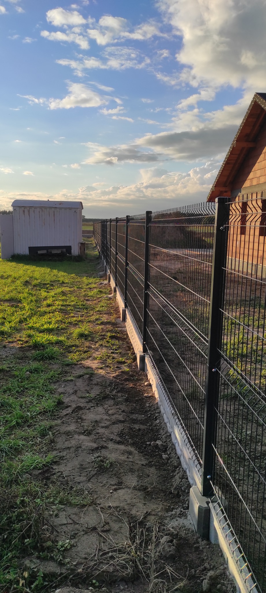 Długi, czarny panel ogrodzeniowy z siatki, osadzony na betonowej podmurówce, rozciąga się wzdłuż trawnika w kierunku białego, blaszanego budynku gospodarczego, z widokiem na zachmurzone niebo...