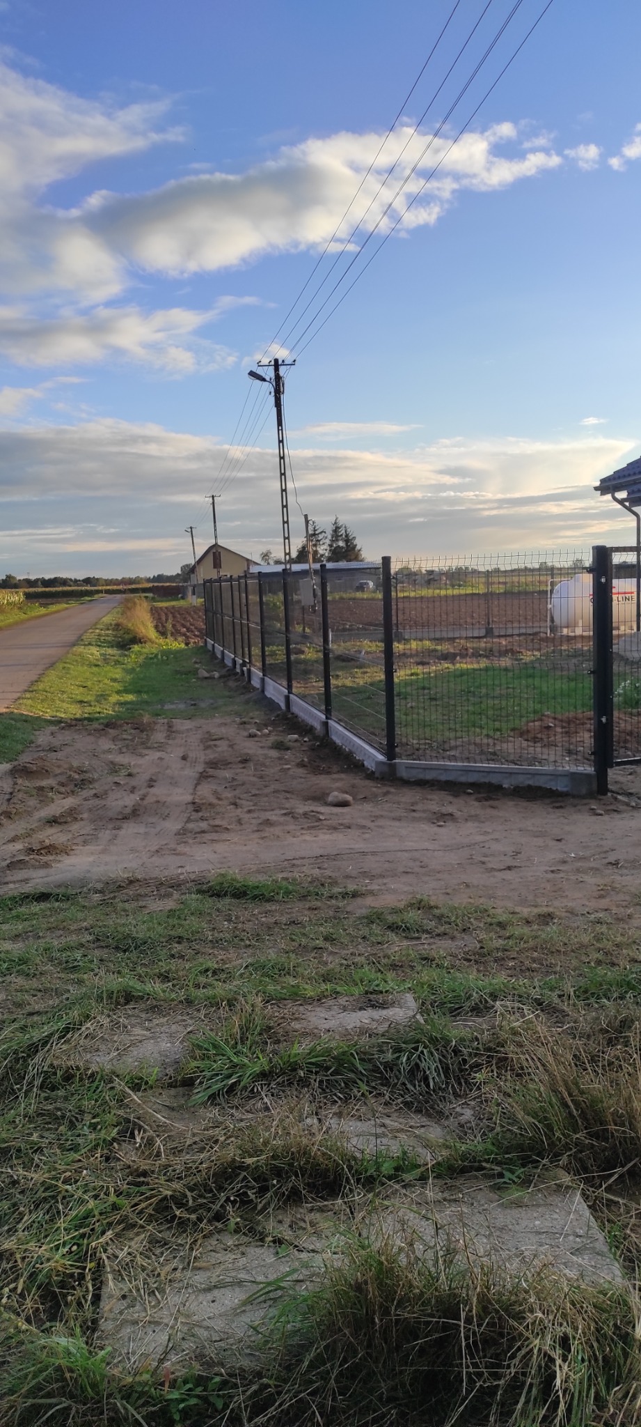 Nowoczesne, czarne ogrodzenie panelowe z podmurówką betonową, widok z perspektywy trawnika w kierunku drogi i zabudowań gospodarczych, błękitne niebo z chmurami w tle.