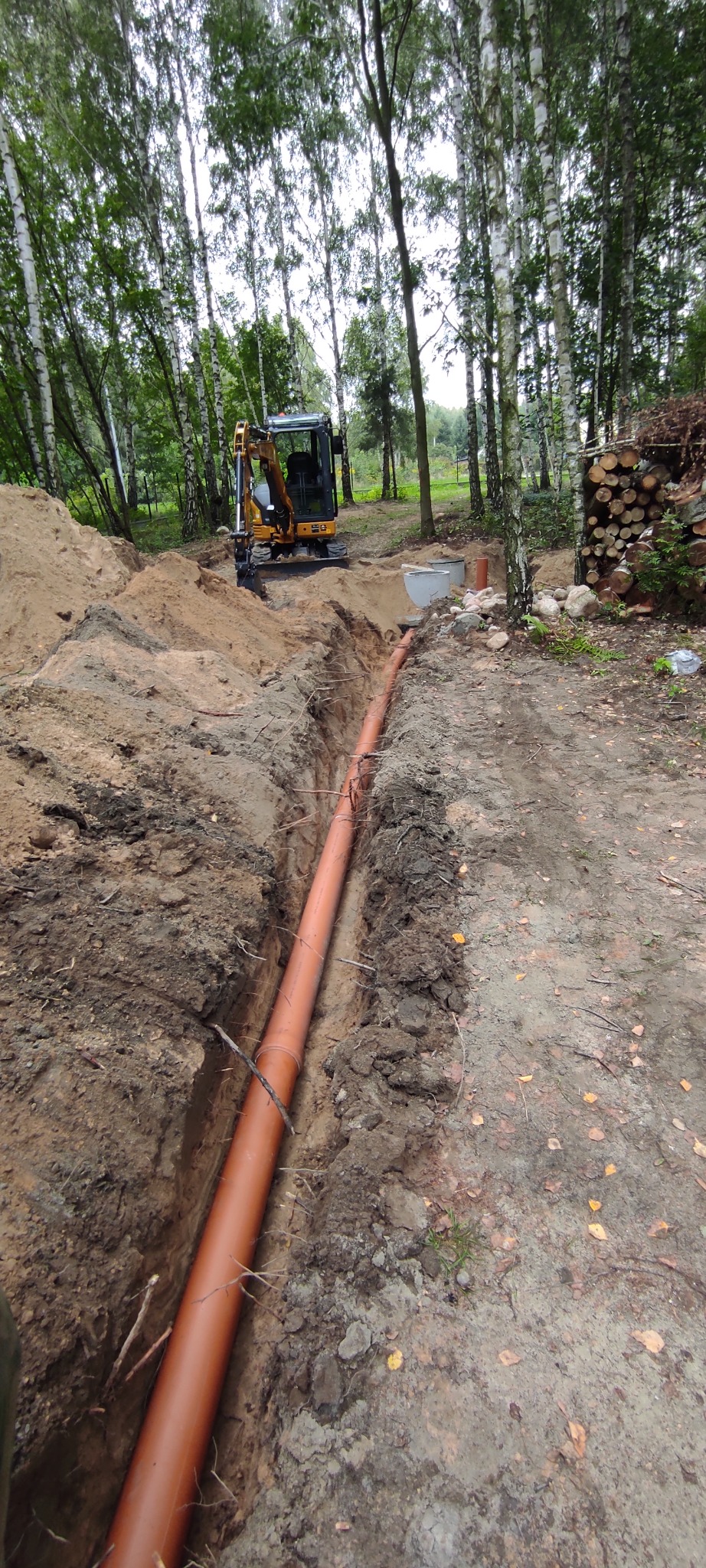 Pomarańczowa rura ułożona w wykopie w ziemi, w tle mała koparka i sterta drewna na tle brzozowego lasu.