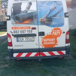 Tył białego samochodu dostawczego Renault z reklamą usług dekarskich i ogrodniczych, w tym zgrzewania papy, z numerem telefonu i tablicą rejestracyjną, zaparkowanego na trawniku.