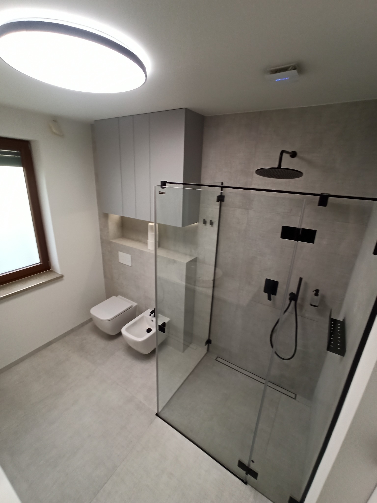 Nowoczesna łazienka w odcieniach szarości z kabiną prysznicową typu walk-in z czarnymi elementami, toaleta i bidet w minimalistycznym stylu, oświetlenie sufitowe LED.