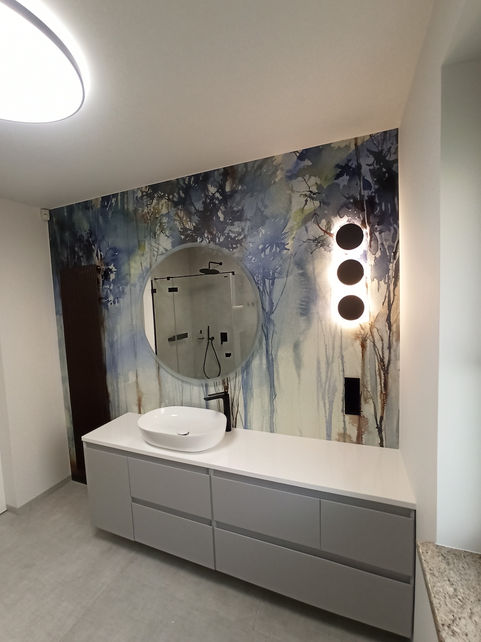 Nowoczesna łazienka z umywalką nablatową, szafką w odcieniach szarości, okrągłym lustrem i dekoracyjną tapetą z motywem leśnym oraz designerskim oświetleniem ściennym.