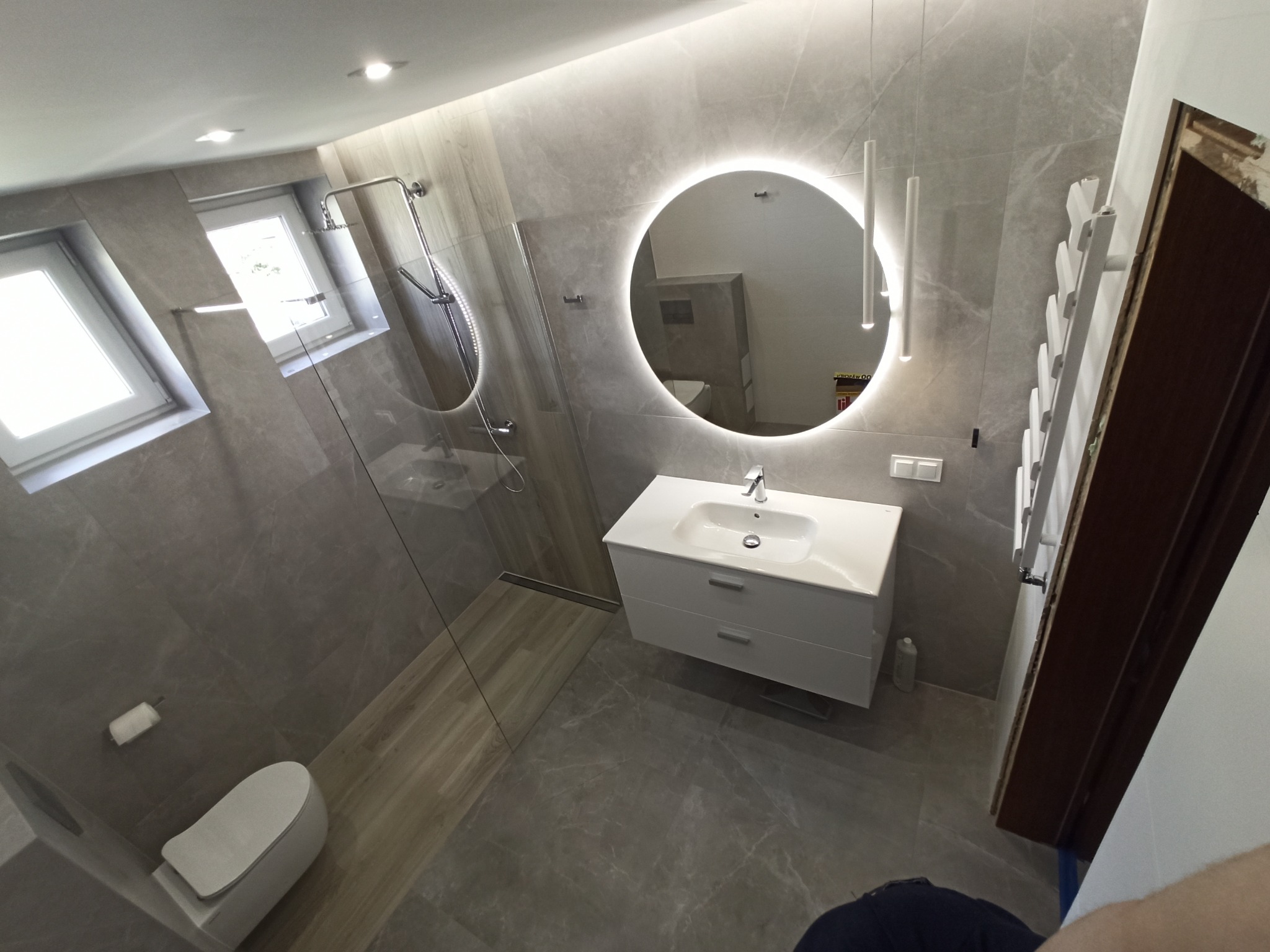 Nowoczesna łazienka w szarościach z okrągłym lustrem podświetlanym LED, kabiną prysznicową typu walk-in i wiszącą toaletą.