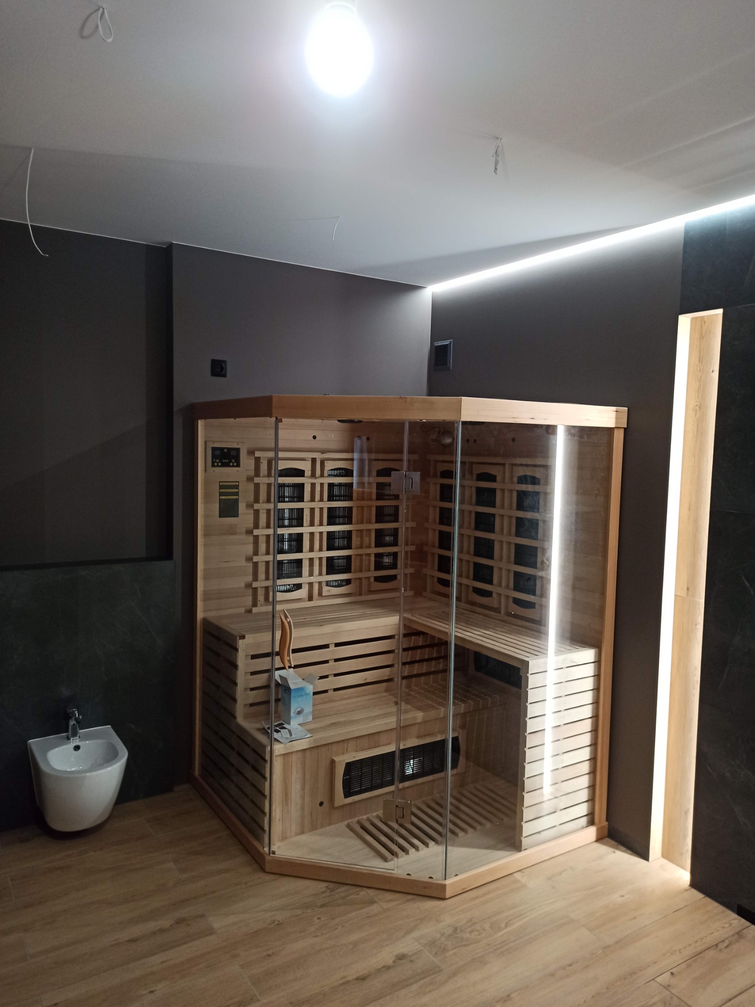 Nowoczesna, narożna sauna infrared z jasnego drewna i szkła, zainstalowana w łazience z beżową podłogą i ciemnymi ścianami, widoczny bidet i niezakończone prace wykończeniowe.