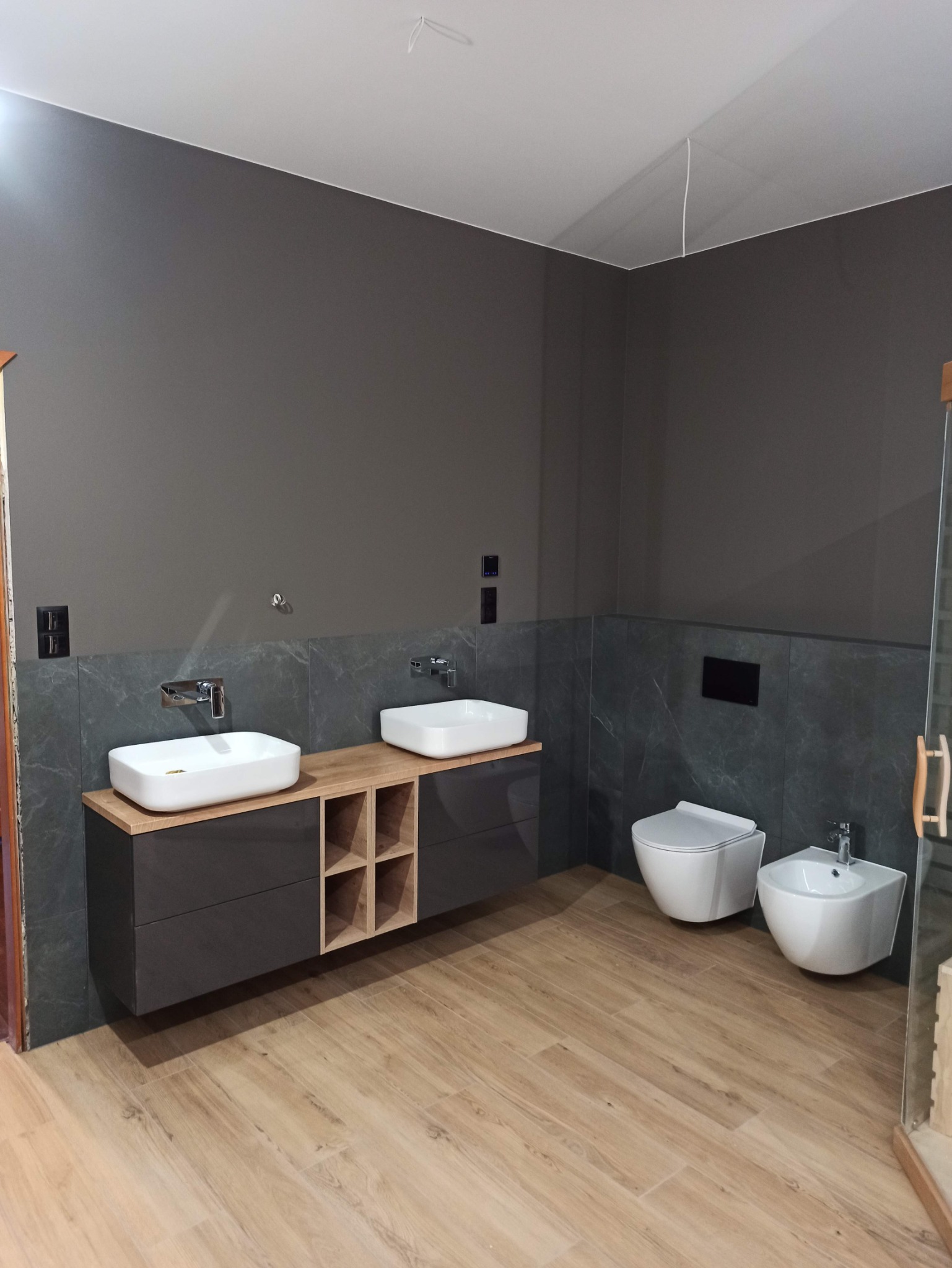 Nowoczesna łazienka z podwójną umywalką nablatową, wiszącą toaletą i bidetem. Szare ściany z marmurowym akcentem, drewnopodobna podłoga. Umywalki na szafce z drewnianym blatem i otwartymi półkami.