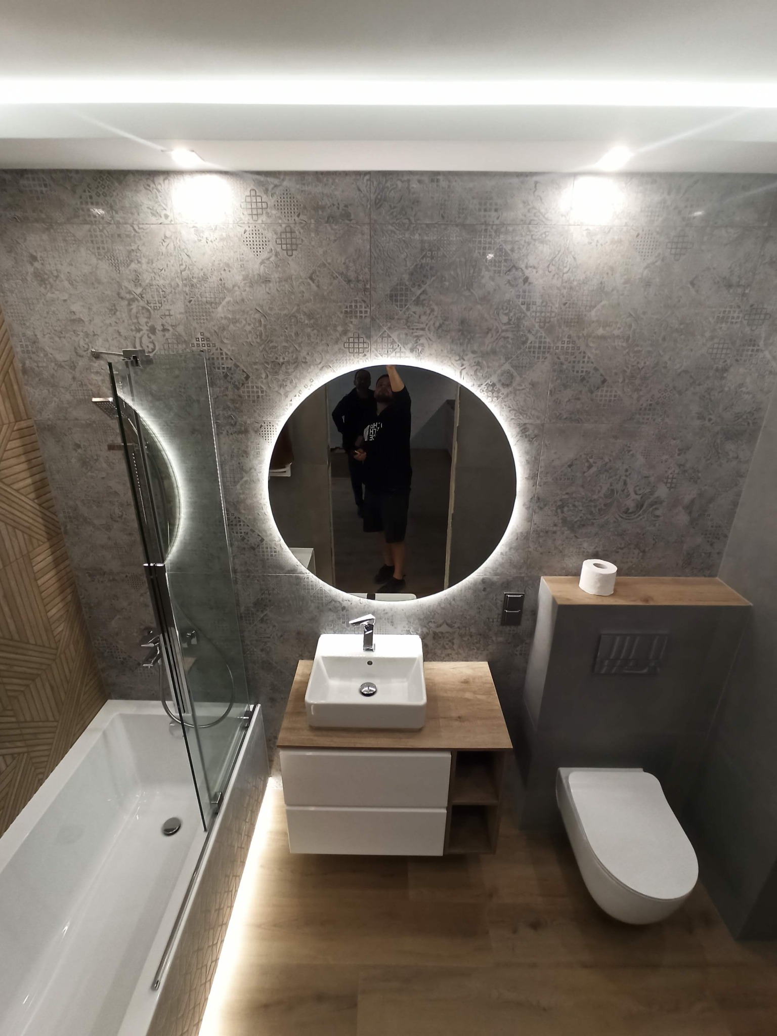 Nowoczesna łazienka z okrągłym lustrem LED, białą armaturą, szarą ceramiką ścienną z wzorami i drewnopodobną podłogą, widok z góry.