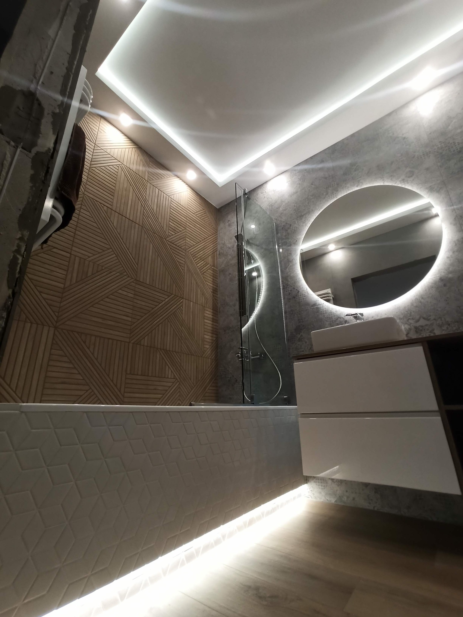 Nowoczesna łazienka z podświetlanym lustrem, drewnianą ścianą z geometrycznym wzorem i podświetlaną wanną wykończoną białymi płytkami o trójwymiarowej strukturze.