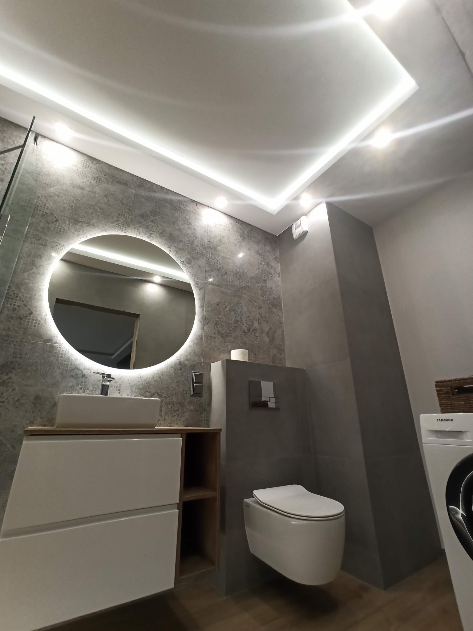 Nowoczesna łazienka w odcieniach szarości z okrągłym lustrem podświetlanym LED, wiszącą toaletą i pralką Samsung, oświetlenie sufitowe LED.