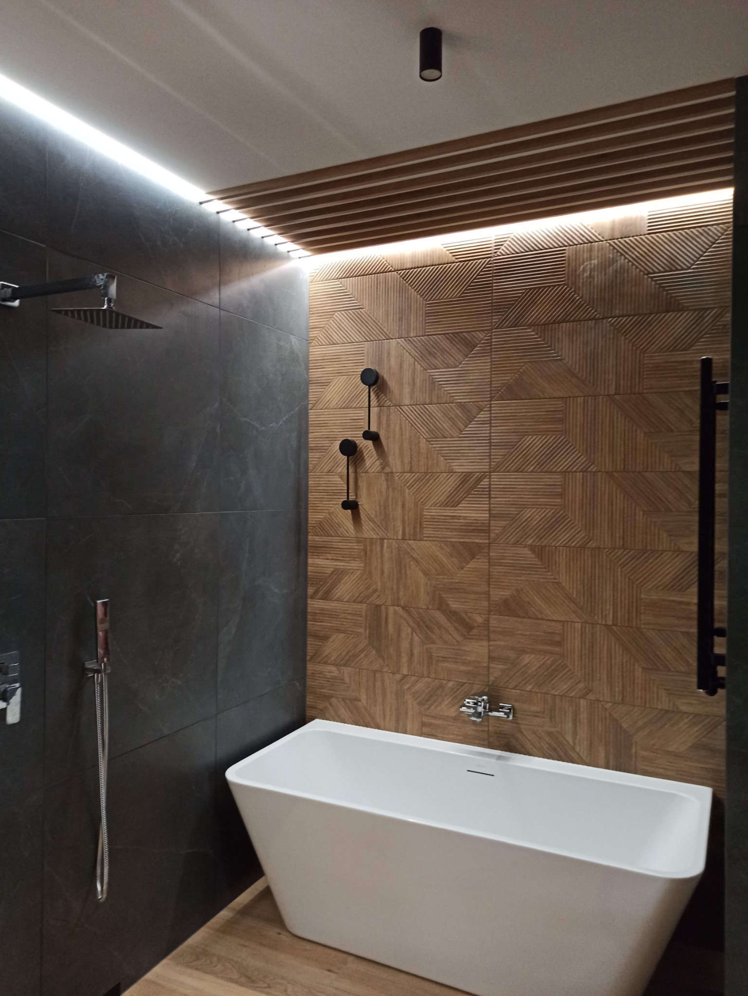 Nowoczesna łazienka z wolnostojącą wanną, deszczownicą, drewnopodobnymi płytkami z geometrycznym wzorem na ścianie i podświetleniem LED pod sufitem.